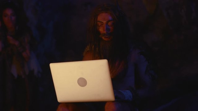 Curious prehistoric indigenous caveman using modern device portable computer in tribe at night. Evolution and technology.