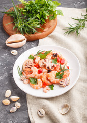 Boiled shrimps or prawns and small octopuses on white ceramic plate on a black concrete background and linen textile. side view.