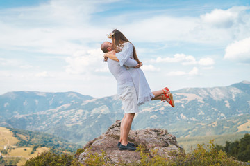 Fototapeta premium Young couple wearing white on the top of the mountain rock man holding his girl friend wife fiancee up hugging and kissing couple in love