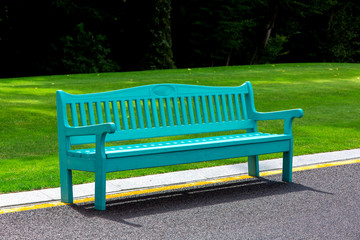 A blue wooden bench lit by sunlight stands on an asphalt road with yellow markings in a park with a green lawn and large trees in the background.