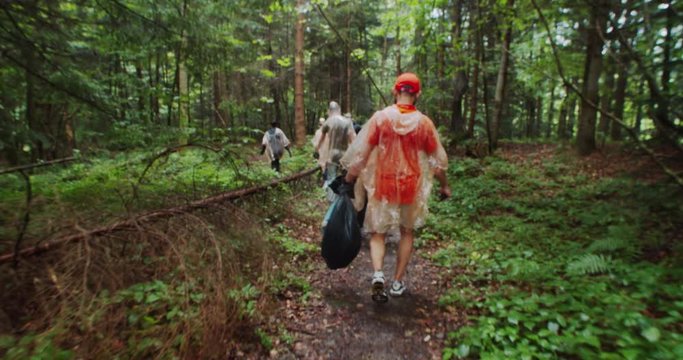 Group of anti-pollution activists in raincoats collecting garbage into bags while walking in the mountain rocky green forest on rainy day. Ecology and environment concept.