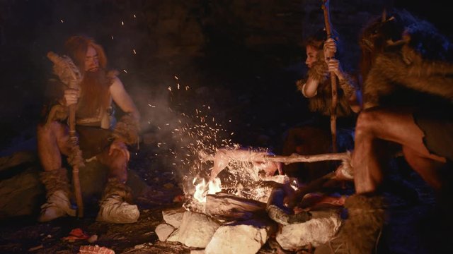 Primal group of neanderthals in animal sking cooking meat sitting at bonfire in cave dwelling. Wild people lifestyle. Back to past.