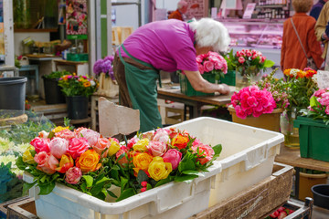 Selected focus at various bouquet of colourful rose and blooming flowers in plastic boxes and blur background of working elder woman florist merchant work at flower stall or outdoor floral shop.