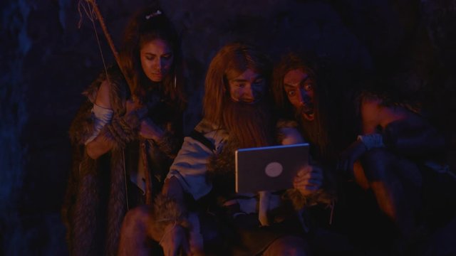 Ancient primitive people family watching videos on modern tablet device outside tribal cave dwelling at bonfire. Evolution of mankind.