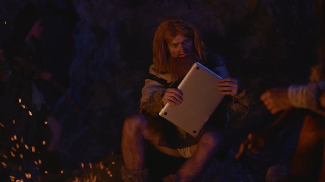 Curious homo sapiens primal tribe leader sniffing looking at modern laptop future device in cave dwelling at bonfire with ancient family. Human evolution.