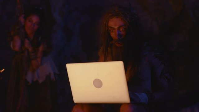 Excited prehistoric homo sapiens looking with surprise on future computer laptop staying at bonfire outside cave. Nomadic primeval humans. Evolution.