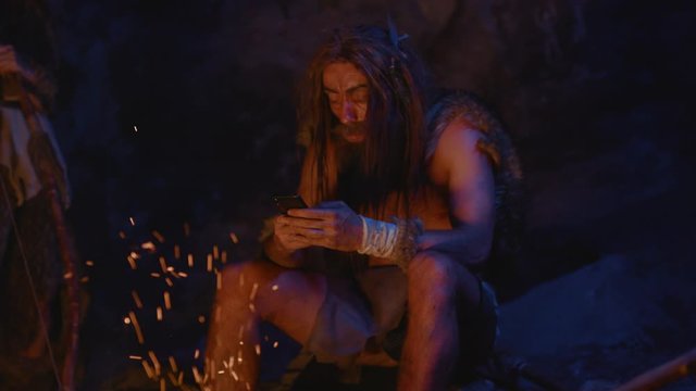 Human ancestor caveman socializing in network smartphone modern gadget near fire. Cro-magnon savage family in animal fur warming up outside cave at night.