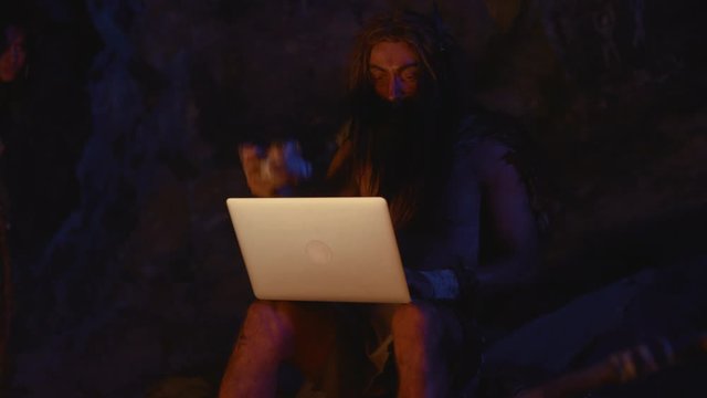 Nomadic cro-magnon caveman in animal fur using future gadget typing on computer keyboard with surprise and curiosity. Evolution concept.