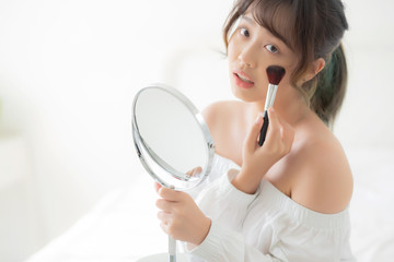 Beauty portrait young asian woman smile with face looking mirror applying makeup with brush cheek in the bedroom, beautiful of girl holding blusher, skin care and cosmetic concept.