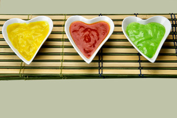 white heart shape bowl with colorful sauces on cork and bamboo mat background