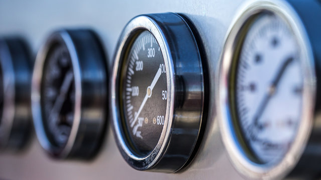 A group of analog measurement gauges on a the side of a fire truck. They are silver, black and white. 