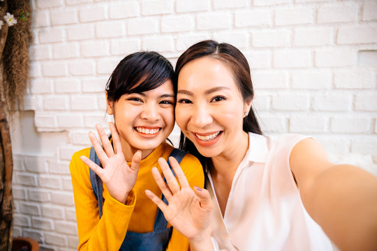 Face of smiling happy Asian teenage daughter and Asian middle-aged mother looking at mobile phone in indoor living room at home. It could be video calling or taking a selfie photo together.
