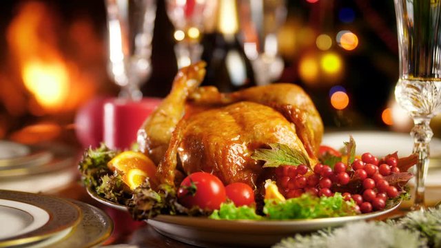Closeup 4k footage of tasty roasted chicken on big plate against burning fireplace and colorful Christmas lights. Dining table served for big family on winter holidays and celebrations.