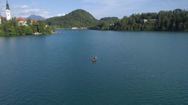 Fly over man sitting in boat at Lake Bled during the day. Tourists visit Bled Island with church dedicated to Assumption of Mary. Mountains and valley at the background.
