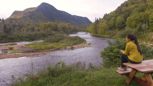Camping nature woman sitting at picnic table enjoying view of wilderness river in Quebec and autumn foliage forest, Canada travel. Parc de la Jacques-Cartier, Quebec.