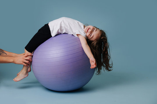 Little girl is relaxing on a yoga ball, while hands holding her over blue