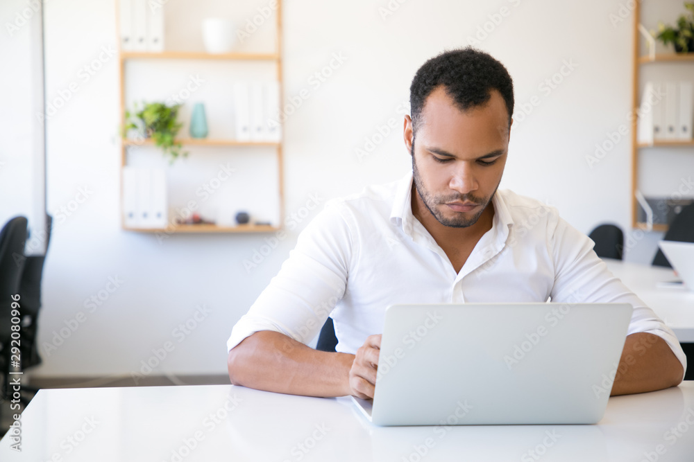 Wall mural serious male professional working on computer at workplace. latin man in white shirt sitting at tabl