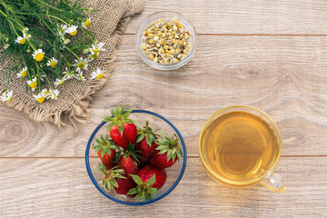 Glass cup of green tea, stawberries with white chamomile flowers on wooden background.