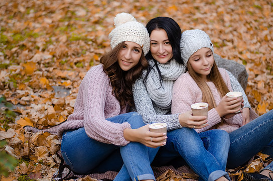 Cute family in a autumn park. Happy mother with two daughters wearing sweaters and jeans, drinking warm coffee or cocoa.  Family sitting on yellow leaves. Golden autumn.