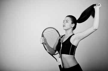 Black and white portrait of beautiful young woman player in sports clothes holding tennis racket while standing against white background.