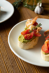 bruschetta with bell shrimp, avocado, pesto sauce and cherry tomato on a white plate on a wooden background. Italian restaurant. Close up food photo