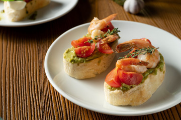 bruschetta with bell shrimp, avocado, pesto sauce and cherry tomato on a white plate on a wooden background. Italian restaurant. Close up food photo
