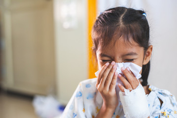 Sick asian child girl who have IV solution bandaged wiping and cleaning nose with tissue on her...