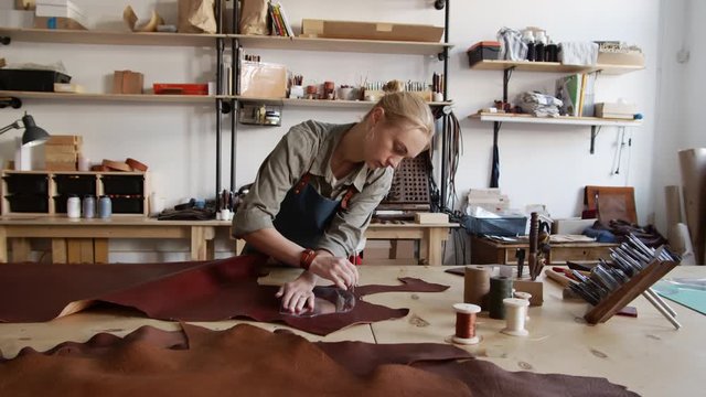 Zoom in of female Caucasian shoemaker circling glass stencil lying on piece of red raw leather with special pen