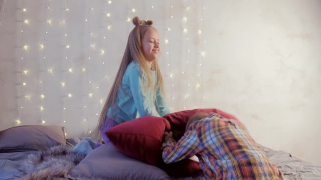 Children cousins teen boy and girl fighting pillows together in bedroom with big window on Christmas holidays. Childhood, friend, friendship, fun concept. New Year interior with garland on wall, xmas.