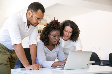 Multiethnic team collaborating on project at one workplace. Business man and women in casual sitting and standing at desk, using laptop, looking at screen and talking. Collaboration concept