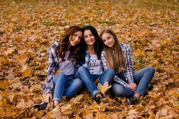 Naklejka na ściany i meble Cute family in a autumn park. Happy mother with two daughters wearing checkered shirts and jeans having fun. Family sitting on yellow leaves. Golden autumn.