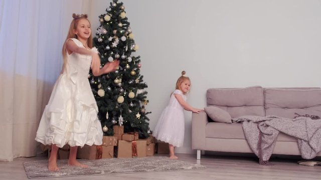 Two girls sisters in beautiful white dresses whirling and dancing near Christmas tree at home in traditional xmas interior in living room. Family time at winter New Year holidays.