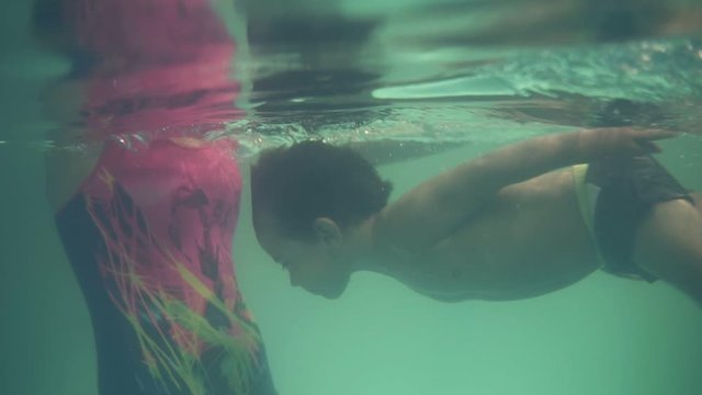 A small swimmer diving into the water and rows with legs under the water, heading to his mother. The boy sits on the edge of the pool, his mother waiting for him in the water, then mother bring him