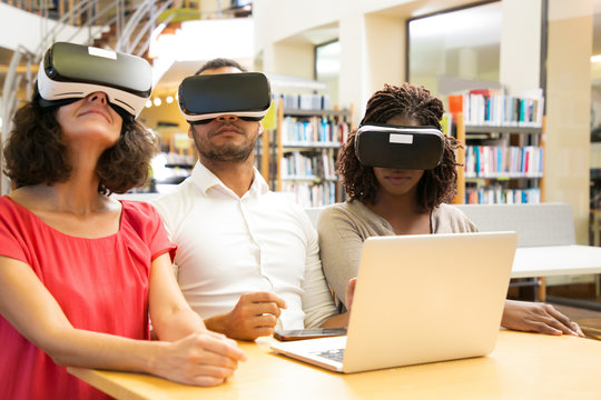 Multiethnic team of adult students enjoying VR experience together. Man and women in virtual reality glasses, sitting at desk with laptop, enjoying experience. Group VR experience concept
