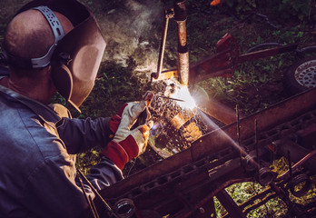 Man using welding machine stick to cut melt iron metal construction outdoors, wearing protective mask and gloves, sparks fly.