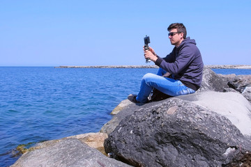 Guy travellet is shooting video os seascape during his vacation. Man tourist starts to record video on smartphone with tripod sits on huge stone at sea. Travel blogger at work in journey. Side view.