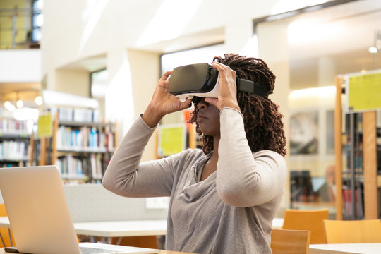 African American female student adjusting VR headset in library. Young black woman sitting at desk with laptop and putting on virtual reality glasses. VR experience concept