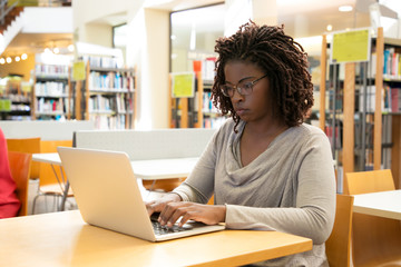 Fototapeta na wymiar Focused female customer using free wi-fi hotspot in public library. Young African American woman sitting at desk and using laptop. Wireless technology concept