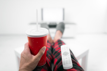 Man drinking soda juice and looking at TV with legs on the table in living room.