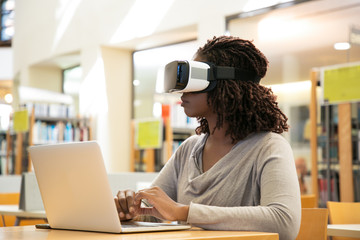 Female library user watching virtual video. Young African American woman wearing virtual reality glasses, sitting at desk with laptop. Innovation for education concept