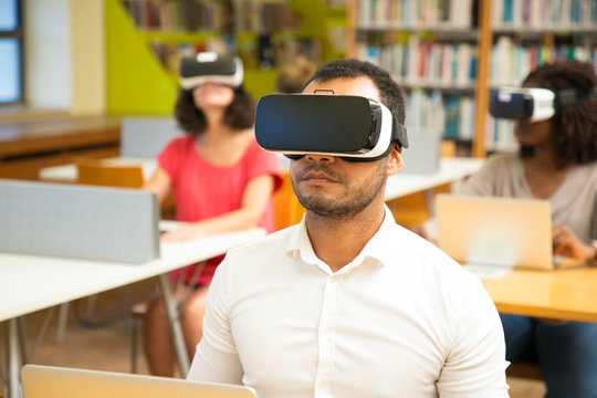 Multiethnic group of students watching virtual presentation in library. Man and women wearing virtual reality headsets, sitting at desks with laptops. Modern computer class concept