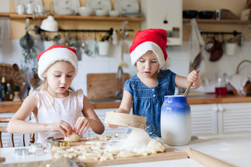 Children are cooking Christmas cookies in cozy home kitchen. Cute kids prepare holiday food for family. Funny little sisters in flour make dough. Lifestyle candid moment. Children chef concept