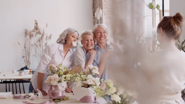 Pan shot of young female florist standing with smartphone in her hands and making photos of three cheerful middle-aged blonde women having fun
