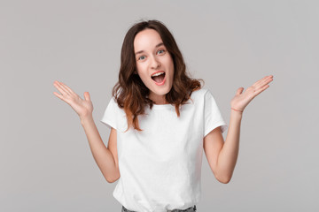 Excited brunette woman in a white tee shrugging her shoulders expressing emotion of surprise standing isolated over grey background. Emotion of surprise.