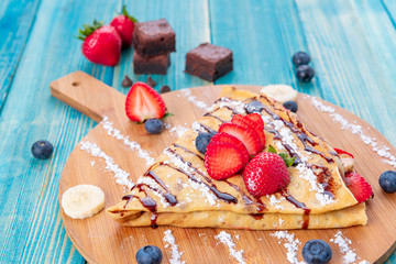 Fototapeta na wymiar Crepe with Strawberries , Berries and Bananas and Brownies - Strawberry Crepe with Fruits with Chocolate Sauce on Top and Sugar Powder on Blue Wood Background