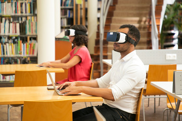 Adult students using VR glasses in public library. Man and woman wearing virtual reality glasses, sitting at desks and using laptop or tablet in computer class. Innovation concept