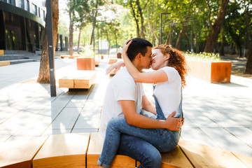 A young couple is sitting on a bench in a modern city, hugging and smiling.