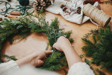Making rustic Christmas wreath. Hands holding fir branches, and pine cones, thread, berries,...