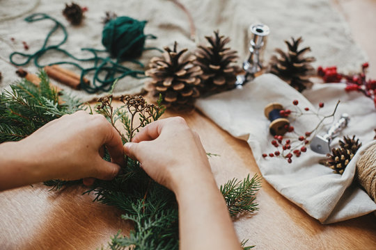 Making rustic Christmas wreath. Hands holding herbs and  fir branches, pine cones, thread, berries, cinnamon on wooden table. Christmas wreath workshop. Authentic stylish still life.