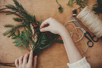 Christmas wreath workshop. Hands holding fir branches, pine cones, berries, thread, scissors on wooden table, flat lay. Making rustic christmas wreath. Authentic rural wreath